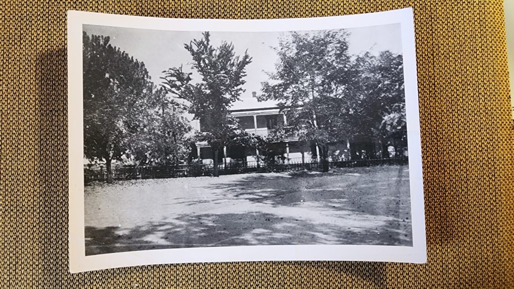 A black and white photo of the old two story house and surrounding areas circa late 1800s.